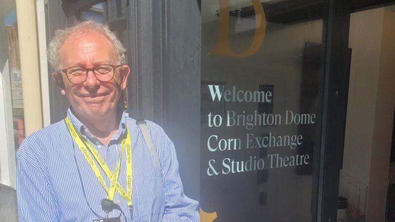 A white man stands outside Brighton Dome waiting to welcome people. Text on the door reads Welcome to Brighton Dome Corn Exchange & Studio Theatre
