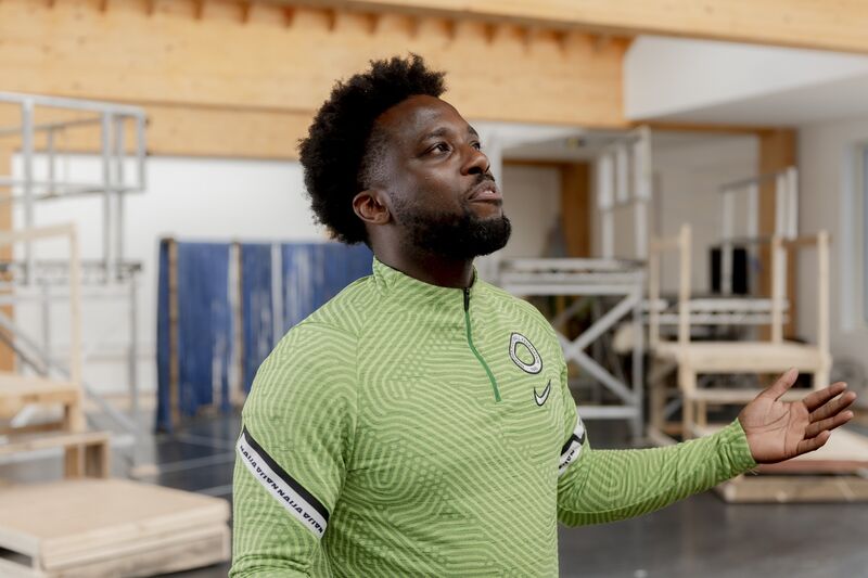 A black man in a green Nigeria football jumper gestures with his left hand, his head is slightly tilted upwards and it looks as though he is giving a speech.