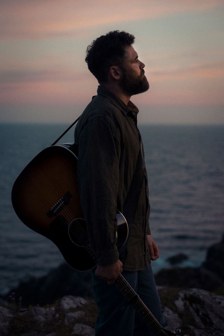 Passenger standing on a cliff in front of a sunset and the ocean. He is looking to the right and holding a guitar under his arm