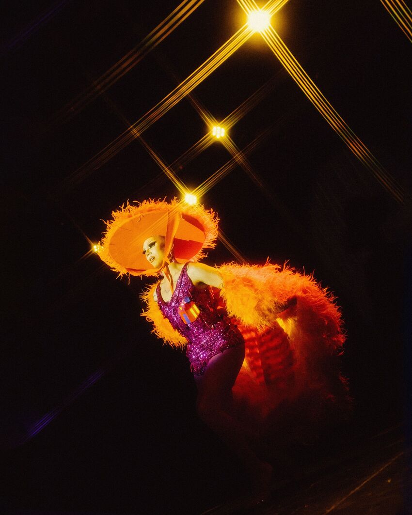 A drag queen in a big orange hat and feather bower plus a sparkly purple dress