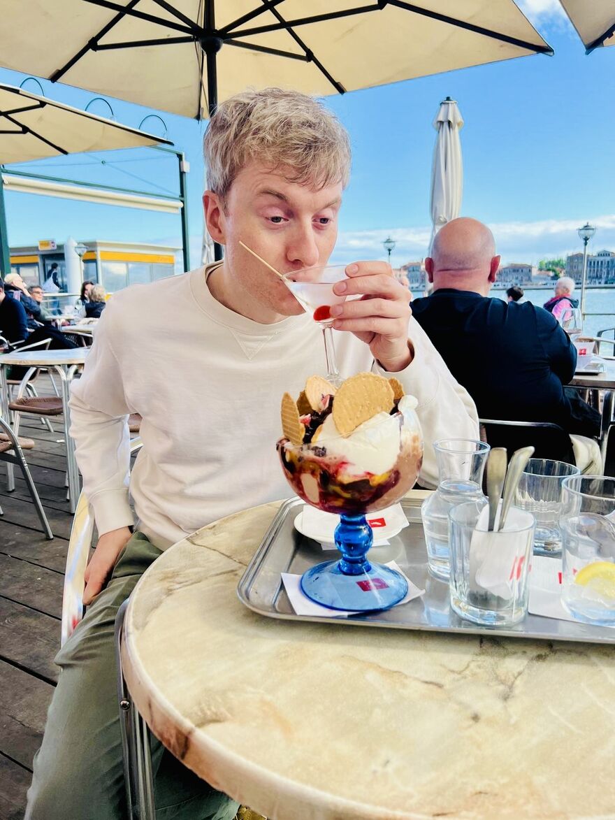 James Acaster sipping a martini at an outside cafe. He has a large sundae in front of him