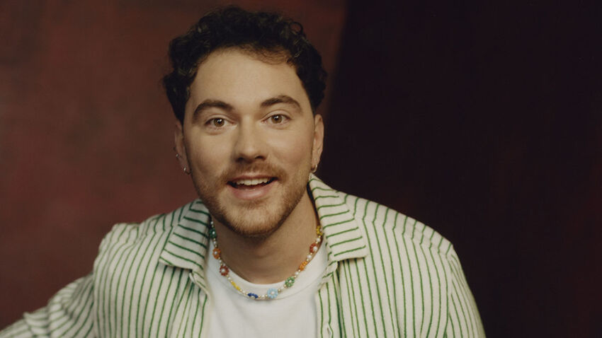 man looks directly forward with a happy expression. He is in a stripey shirt and a plain white t-shirt.