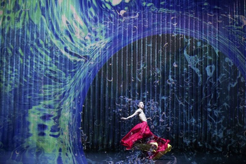 A dancer in flowing red trousers swishes underneath a projection of a wave