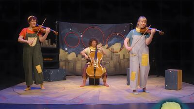 Three people on stage playing string instruments. Two are playing violins and one is playing a cello. They are wearing overalls with patches on.