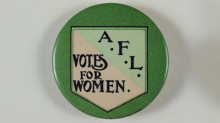 A badge of a light green and tan shield on a dark green background. The badge reads A.F.L Votes for Women