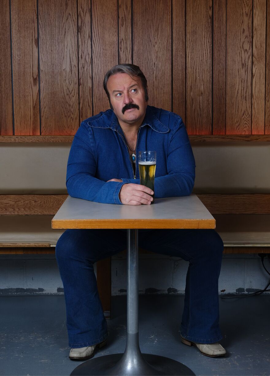 A white man with a moustache wearing blue denim sits at a table holding a beer while glancing quizzically to the top right corner