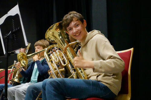 A child holding a French horn smiles and gives the camera a thumbs up