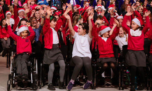 A group of children dresses in bright red school uniforms and Christmas hats are singing as part of a choir. They have their arms in the air and are having a lot of fun