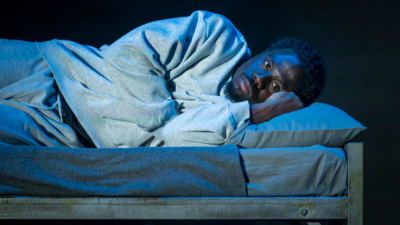 A man in a grey tracksuit lies on his side on a small bed