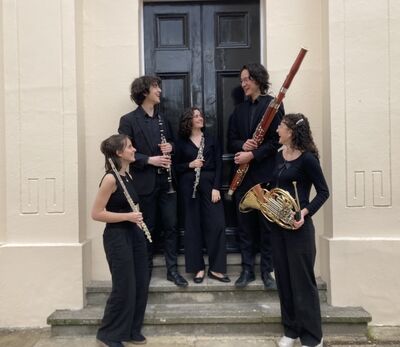 Five people standing in black holding a mixture of wind instruments. They are all smiling at each other