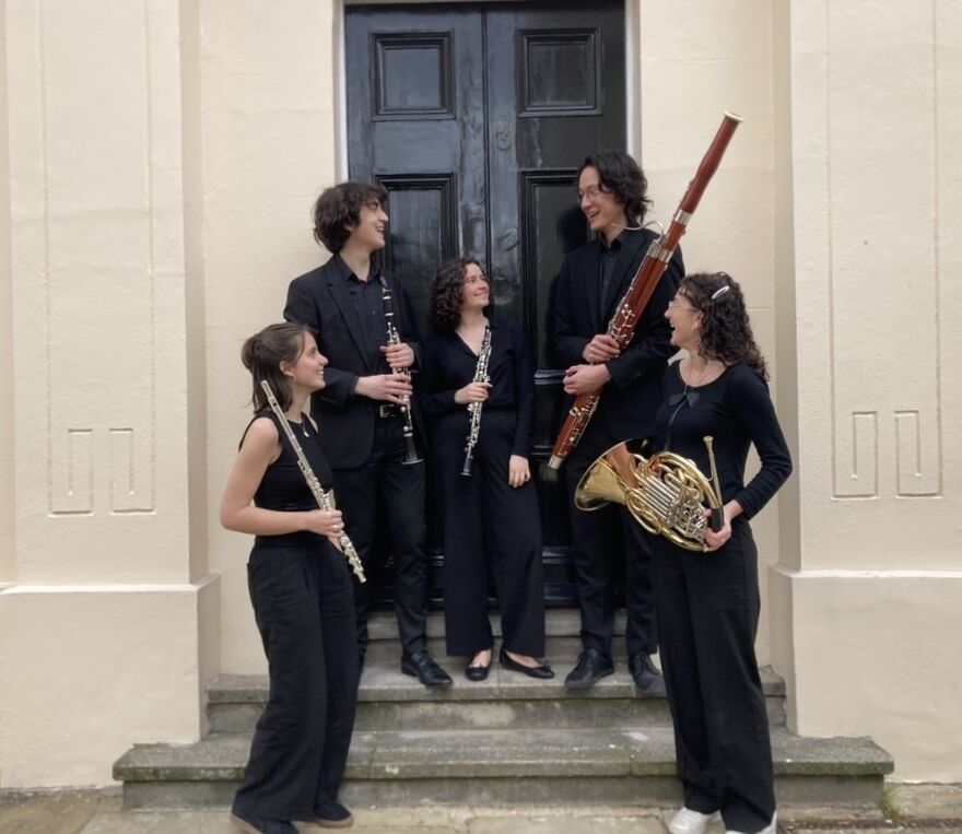 Five people standing in black holding a mixture of wind instruments. They are all smiling at each other