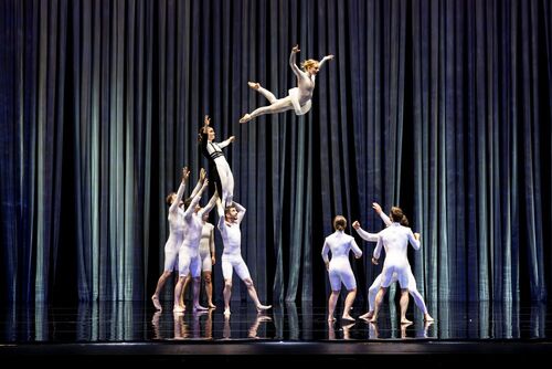 A group of contemporary dancers performs on a theater stage beneath tall, flowing blue-gray curtains. At center, one dancer in a white costume is captured midair in a dramatic leap after being tossed by teammates below, while another performer reaches upward beneath them. Additional dancers in fitted white outfits stand to the right with their arms raised, ready to catch or support. The glossy black stage floor reflects the performers and curtains, emphasizing the height and energy of the acrobatic movement.
