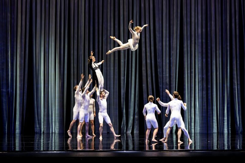 A group of contemporary dancers performs on a theater stage beneath tall, flowing blue-gray curtains. At center, one dancer in a white costume is captured midair in a dramatic leap after being tossed by teammates below, while another performer reaches upward beneath them. Additional dancers in fitted white outfits stand to the right with their arms raised, ready to catch or support. The glossy black stage floor reflects the performers and curtains, emphasizing the height and energy of the acrobatic movement.