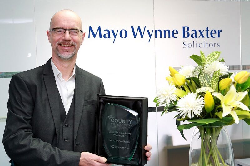 A white bald man with a grey beard holds a clear glass award in a black case. He wears glasses and a black suit.