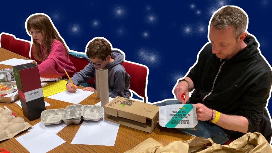 Two children and an adult making crafts at a table. One child is drawing and the adult is cutting a cardboard box. The image is cut out over a blue background with sparkles