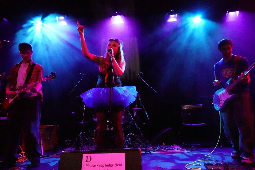Three people on stage performing. Two are playing guitars, one is singing in the middle in a tutu. The stage is dim with purple and blue lights 