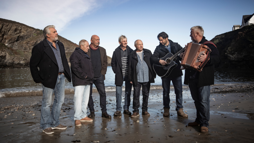 seven men stand together in a group on a beach with dark wet sand. 
