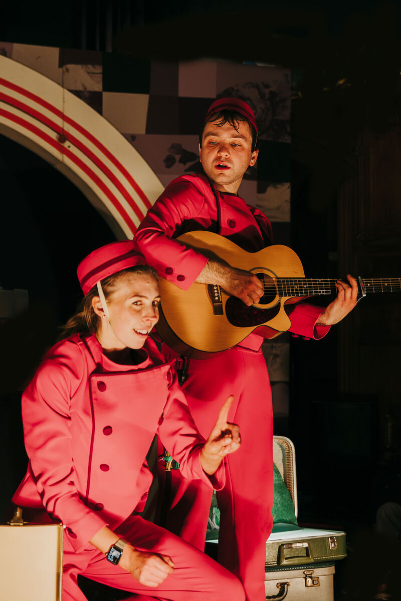 A man in a red jumpsuit and hat plays an acoustic guitar. A woman in a matching outfit is crouched in front of him, looking at the camera and holding up her index finger. 