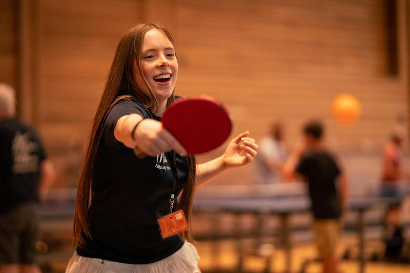 A girl holding a table tennis bat leaning forward like she's about to take a shot. She is smiling
