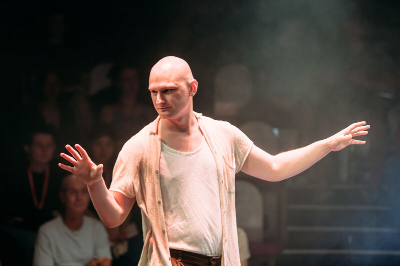 The performer stands in the middle of an in-the-round stage, with haze and ambient white lighting. He is white and bald, and the audience is sitting around him looking at his face