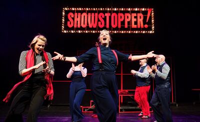 A woman in all black stands centrally, arms outstretched and mouth open, singing. Around her are some other people all wearing black, white and red outfits. Behind them is a red sign that reads SHOWSTOPPER!