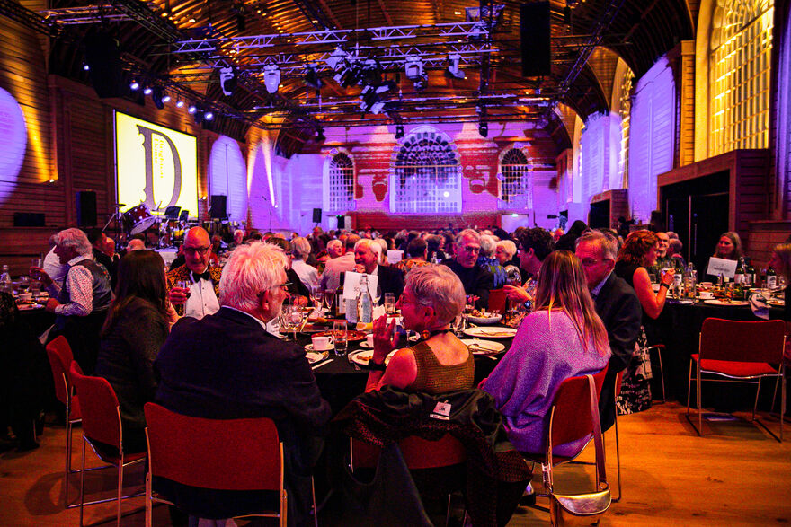 People sat round a round table at a Fundraiser dinner in the Brighton Dome Corn Exchange, that is lit with purple lights