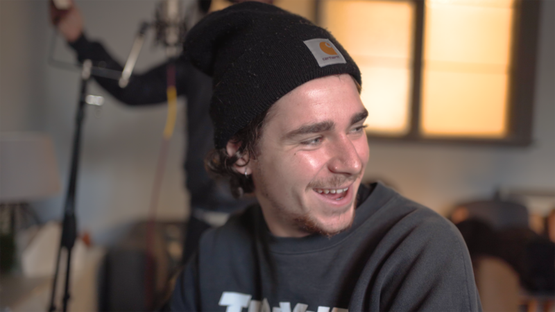 A young man with a beenie wearing a black beenie hat smiles whilst making music
