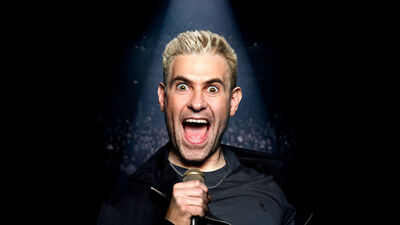 Simon Brodkin holding a microphone with his mouth open under a spotlight from the shoulders up