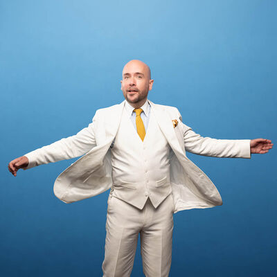 Tom Allen wearing a white suit and yellow tie standing with his arms up by his side. He is smiling at the camera and standing in front of a blue background