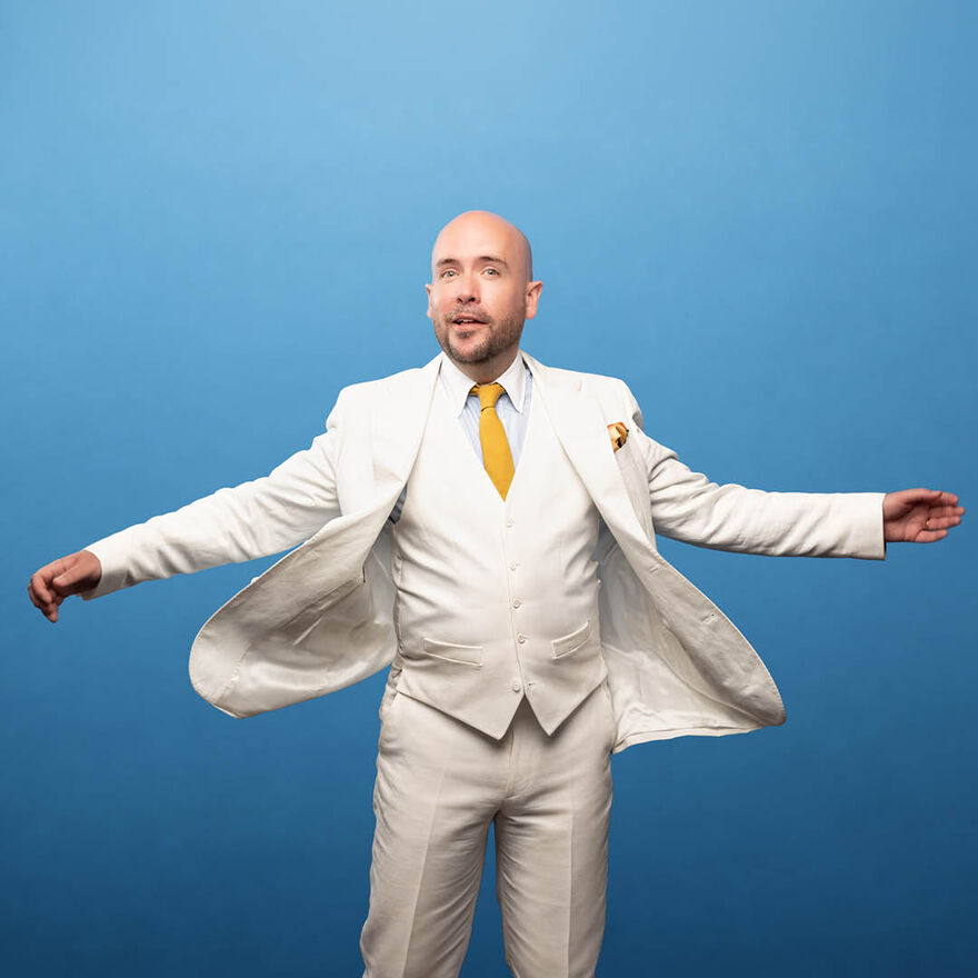 Tom Allen wearing a white suit and yellow tie standing with his arms up by his side. He is smiling at the camera and standing in front of a blue background