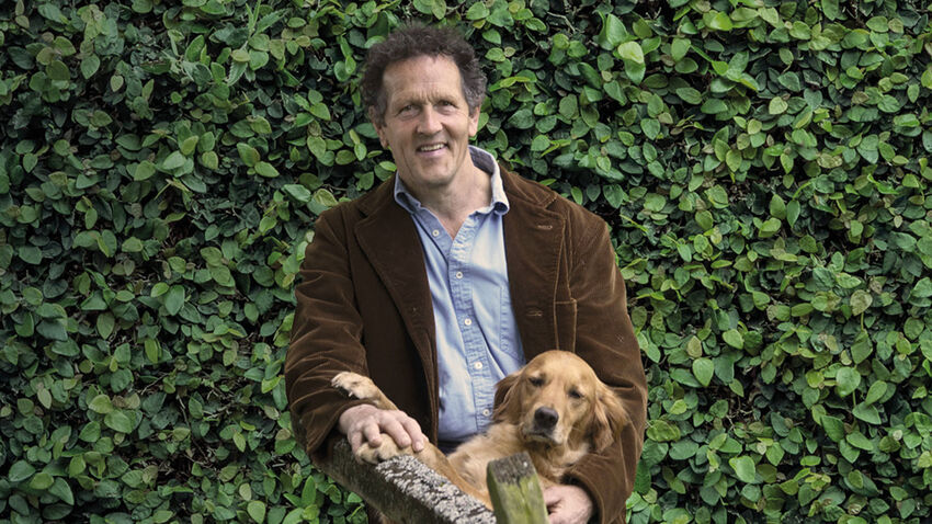 Monty Don standing in front of a green bush, with a dog 