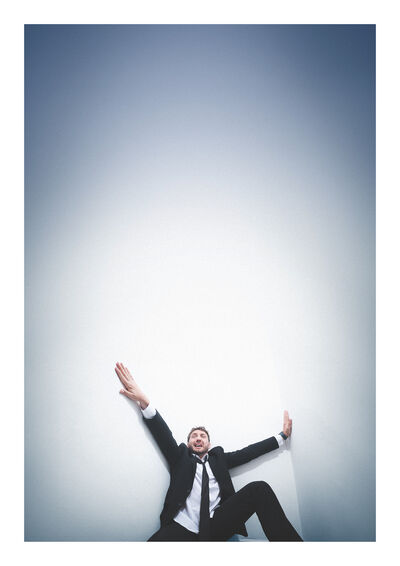 Seann Walsh leaning back against a white wall. He has his arms up and has a confused, stressed facial expression. He looks almost like he is falling down