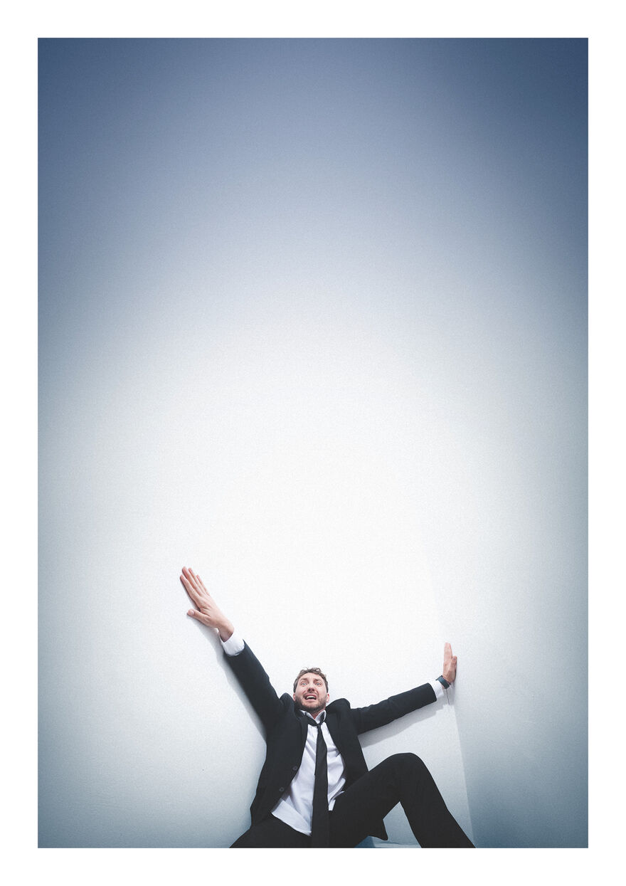 Seann Walsh leaning back against a white wall. He has his arms up and has a confused, stressed facial expression. He looks almost like he is falling down
