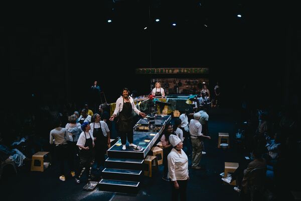 Actors dressed as chefs and cooks standing around a thin stage, surrounded by an audience. The lights are dim, and there is one actor in the centre stage smiling. 