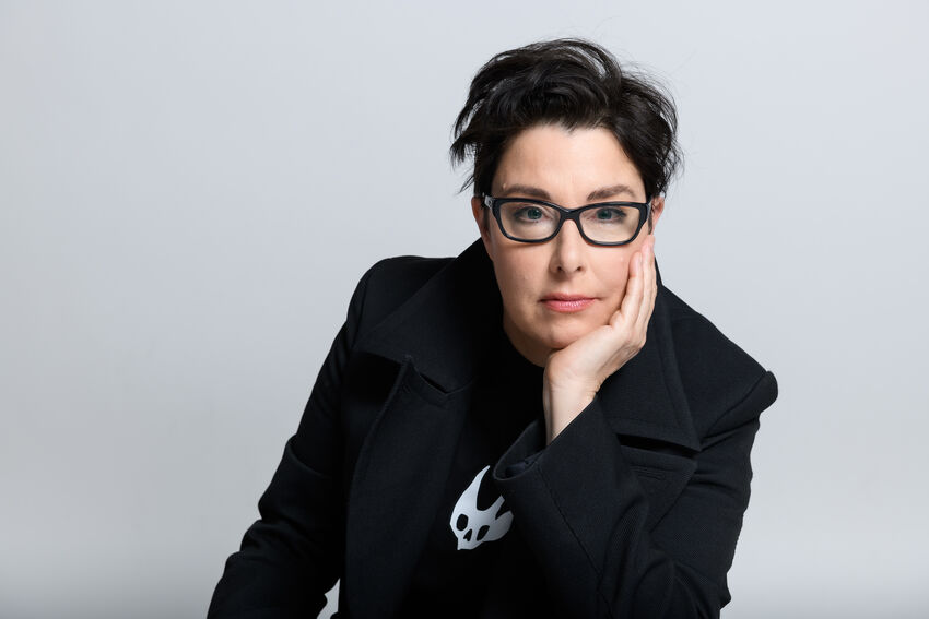 Sue Perkins in a dark suit with her chin resting on her hand