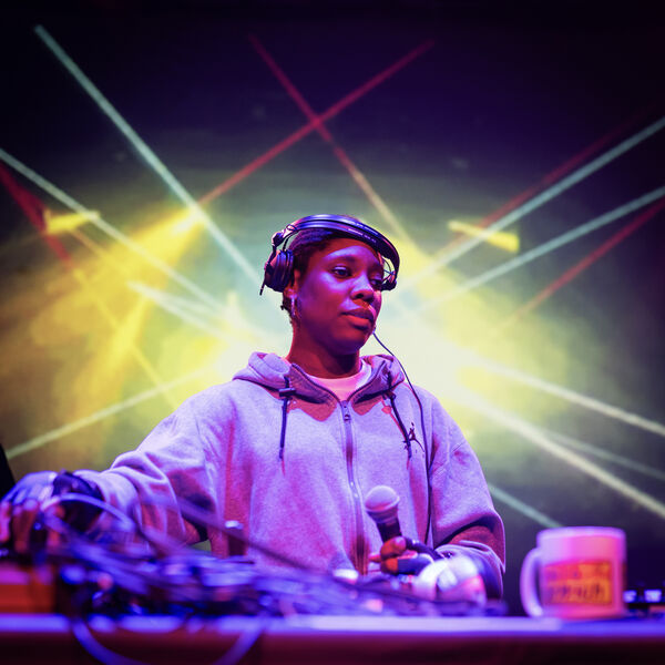 Person DJing and holding a microphone, they are standing in front of yellow, blue and red lazers and smoke