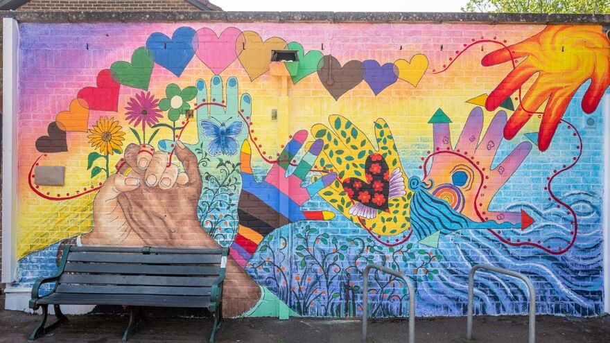 A colourful mural is painted on a wall. There are lots of hands and hearts adorning a background which echoes a sunrise. In front of the wall is a bench and a bike rack
