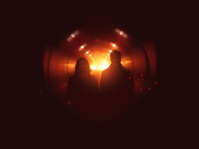 Two people walk side by side through a dark tunnel, silhouetted against a bright orange glow at the far end. Warm overhead lights line the tunnel, and floating sparks or embers create a dramatic, cinematic atmosphere.
