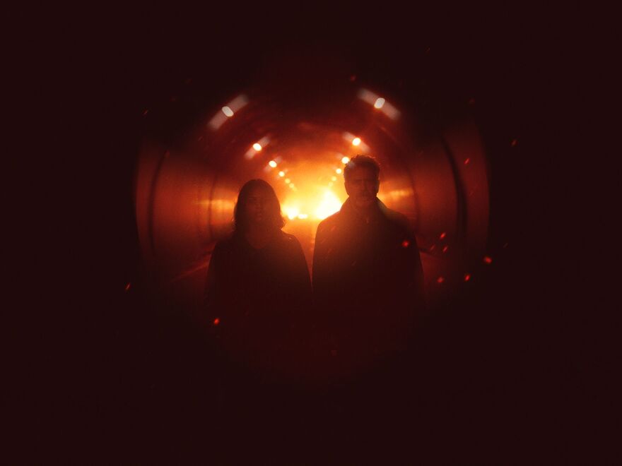 Two people walk side by side through a dark tunnel, silhouetted against a bright orange glow at the far end. Warm overhead lights line the tunnel, and floating sparks or embers create a dramatic, cinematic atmosphere.