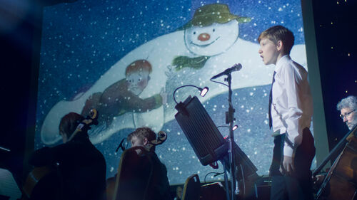 A young boy in shirt and tie sings into a microphone in front of The Snowman film