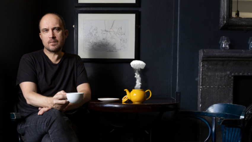 Chris Thorpe sits at a table with his leg crossed, holding a teacup