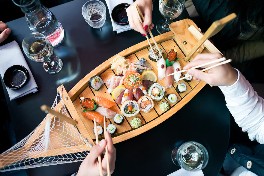A small wooden boat with net sails sits on a black table. It is filled with sushi and people are reaching in with chopsticks in their hands to grab the sushi.
