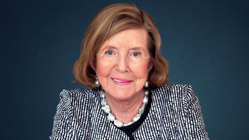 Lady Glenconner smiling in front of a dark blue background, wearing a smart black and white stripe top with a pearl necklace and earrings