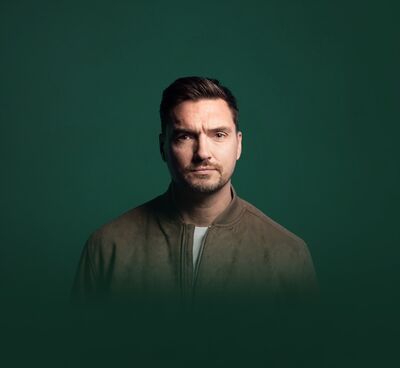 Jack Skipper wearing a suede jacket looking at the camera. He is over a dark green background