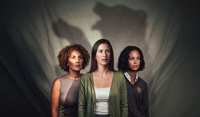 Three people stand shoulder to shoulder in front of a gray backdrop, looking ahead with serious, concerned expressions. Behind them, dramatic shadows form the silhouette of a bear, creating a tense, symbolic atmosphere that suggests hidden fears or looming threats.