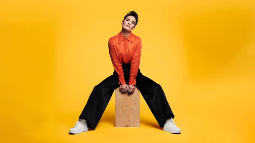 Jen Brister posing on a wooden block in an orange shirt against a yellow backdrop