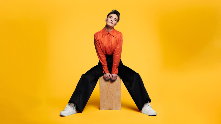 Jen Brister posing on a wooden block in an orange shirt against a yellow backdrop