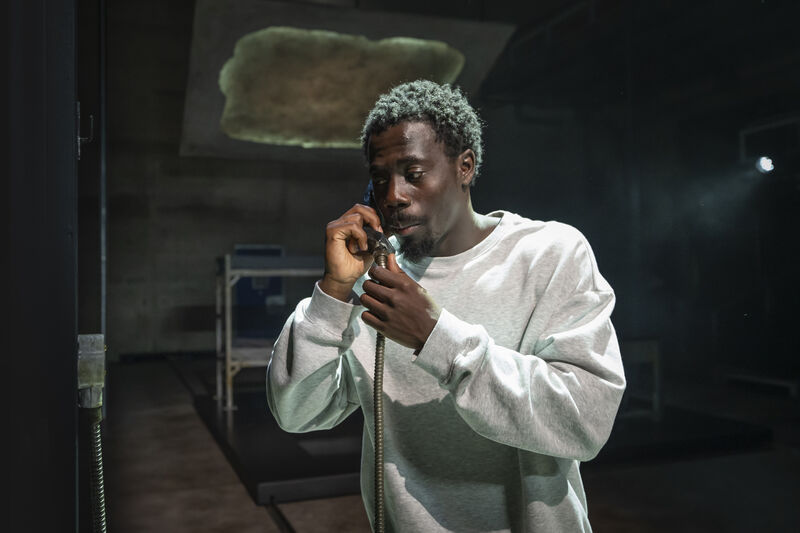 A man wearing a white jumper holding up a phone to his ear. He is standing in a dimly lit room