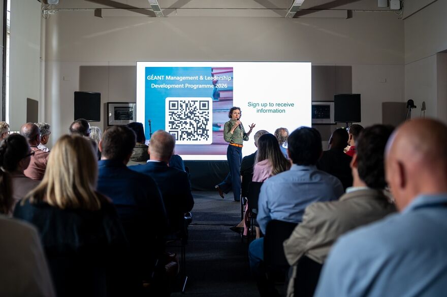 A woman speaks on a microphone, gesticulating with her hands, in front of a seated audience