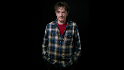 Dylan Moran wearing a blue and white checkered shirt with a red tshirt underneath standing with his hands in his pockets, looking away from the camera with a black backgroud
